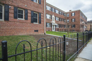 A red brick apartment building with black windows and a black iron fence in front. The grass yard is partially bare, and the sky appears overcast, typical of DC apartments. | DC Apartments by WC Smith