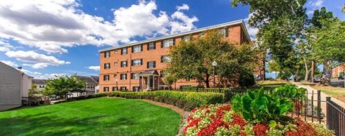 A three-story brick apartment building with a manicured lawn and colorful flower bed in the foreground on a sunny day. These DC apartments offer charm and tranquility amidst bustling city life. | DC Apartments by WC Smith
