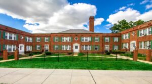 A two-story red brick apartment building with white framed windows, a tall chimney, and a manicured lawn enclosed by a black iron fence under a partly cloudy blue sky, exemplifying the charm of DC apartments. | DC Apartments by WC Smith