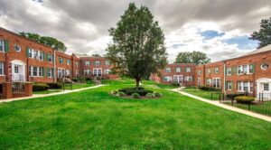 A courtyard with a large tree in the center is surrounded by brick DC apartments with green lawns and pathways. The sky is partly cloudy. | DC Apartments by WC Smith