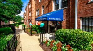 Entrance to an apartment rental office with a blue awning, balloons, and neatly trimmed hedges beside a walkway in a well-maintained brick building complex of DC apartments. | DC Apartments by WC Smith