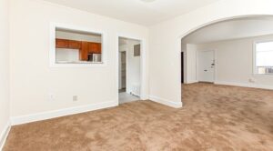 A spacious living room with brown carpet, white walls, a large arched doorway, a window, and a view into a kitchen area through a small interior window—perfect for those seeking DC apartments. | DC Apartments by WC Smith