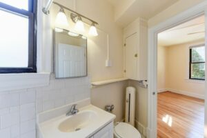 bathroom with sink, toilet, linen cabinet window and view of bedroom at the klingle apartments in cleveland park washington dc