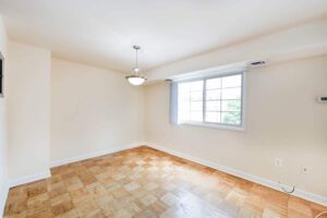 A bright, empty room with a large window, light-colored walls, a wooden parquet floor, and a ceiling light fixture—a perfect example of the modern elegance found in DC apartments. | DC Apartments by WC Smith