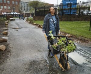 A person pushes a wheelbarrow filled with plants along a paved path on a cloudy day. In the background, others are visible working outside apartments in DC. | DC Apartments by WC Smith