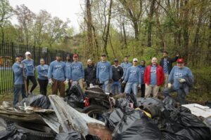 A group of people stands in a wooded area, posing behind a large pile of collected trash bags and debris, indicating participation in a community cleanup effort organized by residents of DC apartments. | DC Apartments by WC Smith