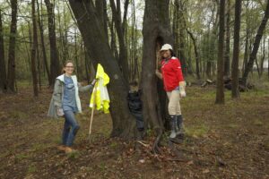 Two people are engaged in a forest activity. One person is holding tools next to a tree with reflective jackets hanging on it, and the other is standing inside a tree hollow. The scene is reminiscent of how DC apartments blend urban life with pockets of lush greenery nearby. | DC Apartments by WC Smith