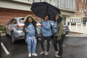 Three women in blue shirts pose with an umbrella in a parking lot of DC apartments. Two stand under the umbrella, while one stands beside them. A car and a large building are visible in the background. | DC Apartments by WC Smith