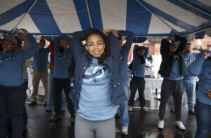 A group of people in blue shirts perform stretching exercises under a large blue and white tent. One woman stands in the front, smiling, as she stretches her arms. The event brings together residents from nearby DC apartments for a community wellness session. | DC Apartments by WC Smith