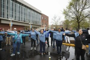 A group of people stands outside a building, performing arm stretches together. Many are wearing blue shirts and jackets. The ground is wet, and trees and other people are visible in the background, possibly near some DC apartments. | DC Apartments by WC Smith