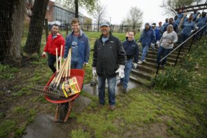 A group of people walks up a grassy hill with gardening tools in a wheelbarrow, participating in an outdoor community service event near DC apartments. | DC Apartments by WC Smith