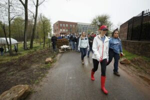 A group of people walk down a paved path outside on a rainy day, with some carrying supplies on a cart. Trees and a building, possibly DC apartments, are visible in the background. | DC Apartments by WC Smith