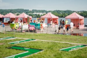 A scene at a lakeside fair with striped red and white tents, various activities, and a mini-golf course. People from nearby DC apartments are walking and engaging in different games on a sunny day. | DC Apartments by WC Smith