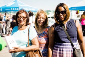 Three women standing outdoors at an event, smiling at the camera. The woman on the left holds a small bag. People are visible in the background near a blue canopy, likely enjoying local amenities close to DC apartments nearby. | DC Apartments by WC Smith