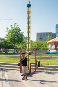 A young boy stands next to a strength tester game at an outdoor fair, holding the mallet. The background includes trees, a bouncy castle, and a building reminiscent of DC apartments. | DC Apartments by WC Smith