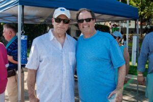 Two men wearing sunglasses, one in a white shirt and hat and the other in a blue shirt, smile for a photo outdoors under a blue canopy at an event for apartments in DC. Other people are gathered in the background. | DC Apartments by WC Smith