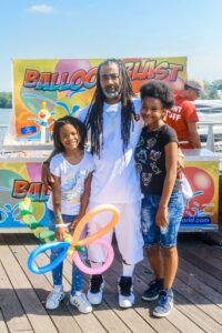 A man stands on a wooden deck with his arms around two smiling young girls. They are in front of a booth labeled "Balloon Blast," with colorful balloons visible. One girl holds a balloon sculpture, enjoying a festive day before heading back to their cozy apartments in DC. | DC Apartments by WC Smith