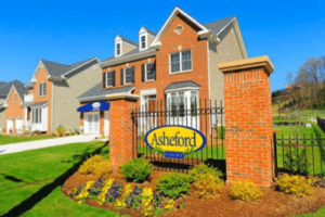 A suburban neighborhood with a brick gate sign reading “Asheford.” The houses are two-storied with gabled roofs, and the area is landscaped with shrubs and plants, offering a peaceful retreat that contrasts the bustling lifestyle of DC apartments. | DC Apartments by WC Smith