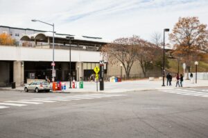 A street scene shows a subway entrance with a sign indicating the letter "M." Nearby, vehicles are parked by a row of newspaper stands, and a few pedestrians walk past. The dynamic city environment perfectly complements the sought-after lifestyle of those residing in nearby apartments in DC. | DC Apartments by WC Smith