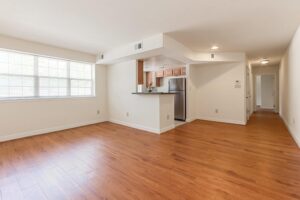 An empty living room with wood flooring, an open kitchen with stainless steel appliances, and a hallway. Large window on the left lets in natural light. Walls are painted in off-white. | DC Apartments by WC Smith