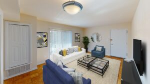 Modern living room with a white sofa, two blue armchairs, a glass coffee table, abstract wall art, and a TV. Beige walls, wooden floor, and a large window with vertical blinds. | DC Apartments by WC Smith