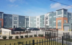 This modern apartment complex, a shining example of prime real estate, boasts a blend of gray and brick facades with large windows. A spacious parking area lies in front, encircled by a black metal fence. It’s no wonder this property has been recognized with Best Deal Awards. | DC Apartments by WC Smith