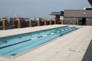 Rooftop swimming pool with lane dividers, surrounded by a deck. City skyline visible in the background under a clear sky. | DC Apartments by WC Smith