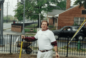 Man holding a rake stands on a lawn near a wheelbarrow. A street and parked SUV are in the background, next to a brick building and fence. | DC Apartments by WC Smith