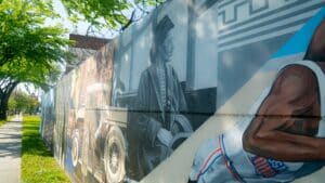 A mural on a wall in Deanwood showcases historical figures, a vintage car, and an athlete in motion. Trees cast shadows on the vibrant art. | DC Apartments by WC Smith