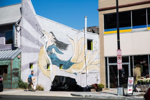 In Glover Park, a street mural showcases a woman with flowing hair and a yellow dress. Nearby, a man strides along the sidewalk beside a parked black vehicle, adding life to the vibrant community scene. | DC Apartments by WC Smith