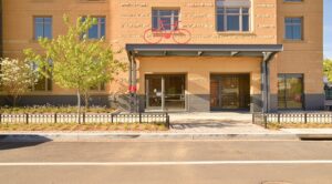 Brick building entrance with a bike symbol above. There are plants and a metal fence in front, and a road in the foreground. | DC Apartments by WC Smith