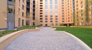 Courtyard of a modern apartment complex with beige brick buildings, a paved walkway, small trees, and a grassy area. | DC Apartments by WC Smith