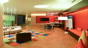 Modern room with colorful decor, including a multicolored rug, white and red seating, a round table with chairs, and a large TV on a wooden wall. Ceiling lights illuminate the space. | DC Apartments by WC Smith