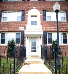 Entrance of a brick apartment building with a white door, black shutters, and a streetlamp on the right. Steps lead up to the door, flanked by black metal railings. Building number 900 is visible. | DC Apartments by WC Smith