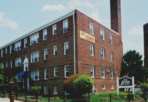 Red-brick apartment building with a "Now Leasing" sign on the wall and a white sign in front reading "Jetu Apartments." Green lawn and fence in the foreground. | DC Apartments by WC Smith