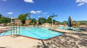 Outdoor swimming pool with lifeguard chair, water features, and striped umbrellas. Surrounding area has trees and buildings under a blue sky. | DC Apartments by WC Smith