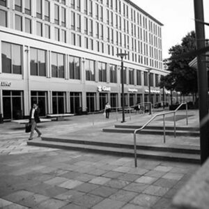 Black and white image of the Van Ness building with large windows and multiple floors. People stroll on a paved area nearby, while stairs with railings stand prominently in the foreground. | DC Apartments by WC Smith