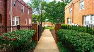 The pathway, flanked by hedges and charming brick buildings under rent control, guides visitors to an open area where trees stand serenely in the background. | DC Apartments by WC Smith