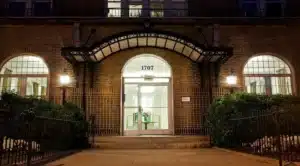 The entrance of the brick building in Washington DC boasts arched windows and is marked 1707. Lit lamps and decorative ironwork above glass doors enhance its charm, while greenery flanks the stairs. Perfect for those seeking cat-friendly apartments with style. | DC Apartments by WC Smith