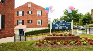 A charming brick building with a sign reading "Worthington Woods" sits amidst vibrant flowers and balloons, offering cat-friendly apartments. Conveniently located near Washington DC, it proudly displays a phone number for inquiries. | DC Apartments by WC Smith