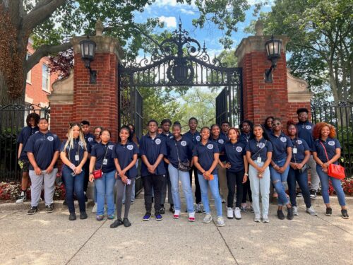 A diverse group of young adults in matching navy shirts, part of the Summer Youth Employment program, stands in front of an ornate iron gate and brick pillars, posing for a group photo outdoors. | DC Apartments by WC Smith