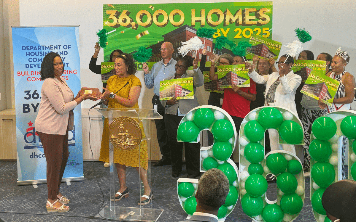 Two women are on stage at the Terrace Manor event, one handing a plaque to the other. In the foreground, a group holds "36,000 Homes by 2025" signs and green balloons, celebrating strides in housing production. | DC Apartments by WC Smith