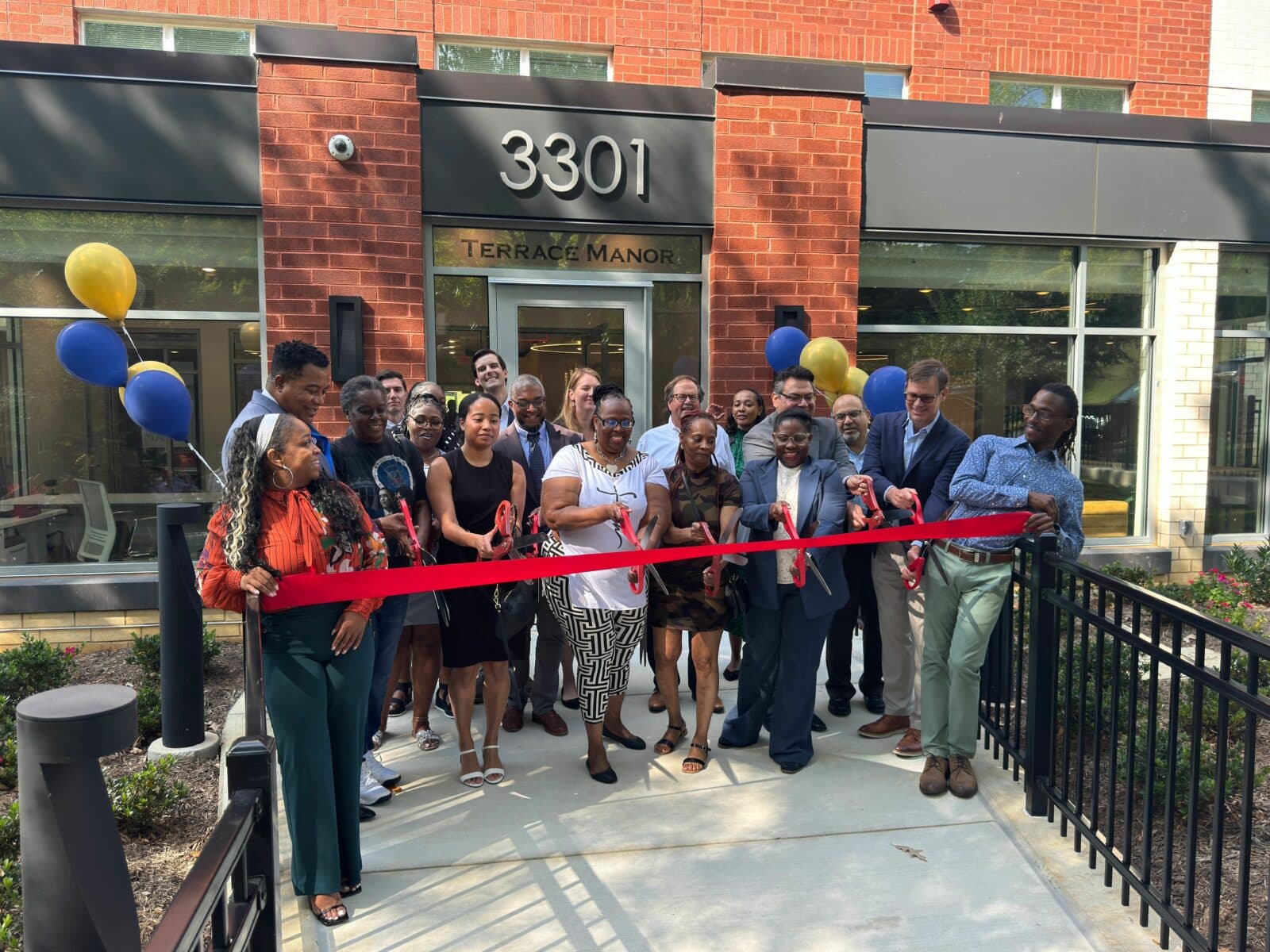 A group of people standing in front of a building labeled "Terrace Manor" participate in a ribbon-cutting ceremony, with the ribbon being cut by a woman in the center. WC Smith representatives are also present to commemorate the event. | DC Apartments by WC Smith