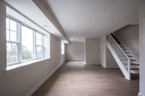 A spacious, empty room with light wood flooring, large windows, and a staircase leading to an upper floor. Walls are painted white, creating a bright atmosphere. | DC Apartments by WC Smith