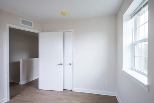 Empty room with light-colored walls, wood flooring, a window on the right, and an open door leading to another space. | DC Apartments by WC Smith