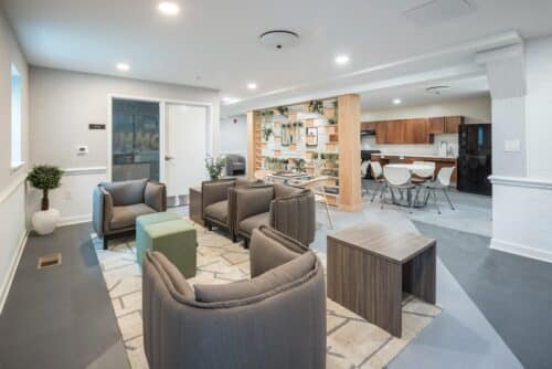 A modern lounge area with gray armchairs, a green ottoman, and a wooden table, adjacent to a kitchen with a dining area, featuring plants and a bookshelf. | DC Apartments by WC Smith