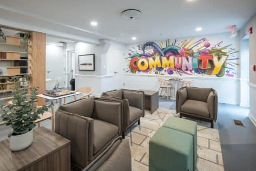 Modern lounge area with cushioned seating, a colorful "Community" mural on the wall, a chessboard, and green potted plants. | DC Apartments by WC Smith