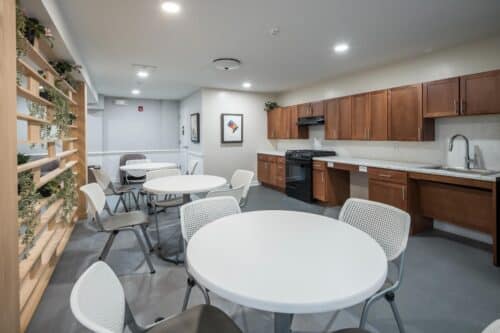 A modern communal kitchen with wooden cabinets, black appliances, round white tables, and gray chairs. Walls have decorative shelves and a framed artwork. | DC Apartments by WC Smith