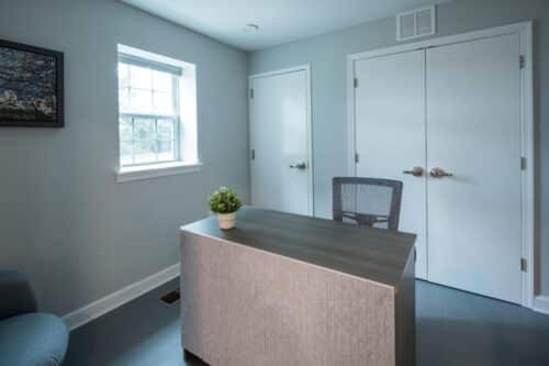 Small office with a desk, chair, and potted plant. A window on the left lets in natural light. Two closed doors are on the right wall. | DC Apartments by WC Smith