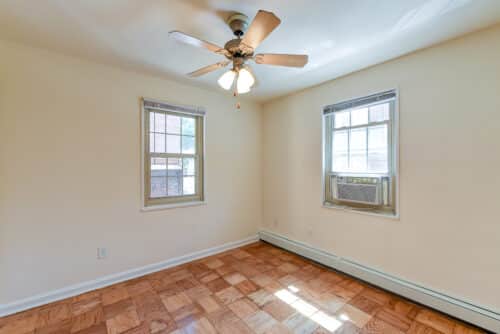 An empty room with wooden floors, two windows, a ceiling fan, and an air conditioning unit installed in one window. | DC Apartments by WC Smith